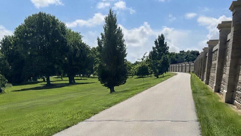 Paved Greenway path in Grand Forks running between the concrete flood wall and open grass with mature trees, with the post title The Grand Forks Greenway Was Never Supposed To Be a Park.