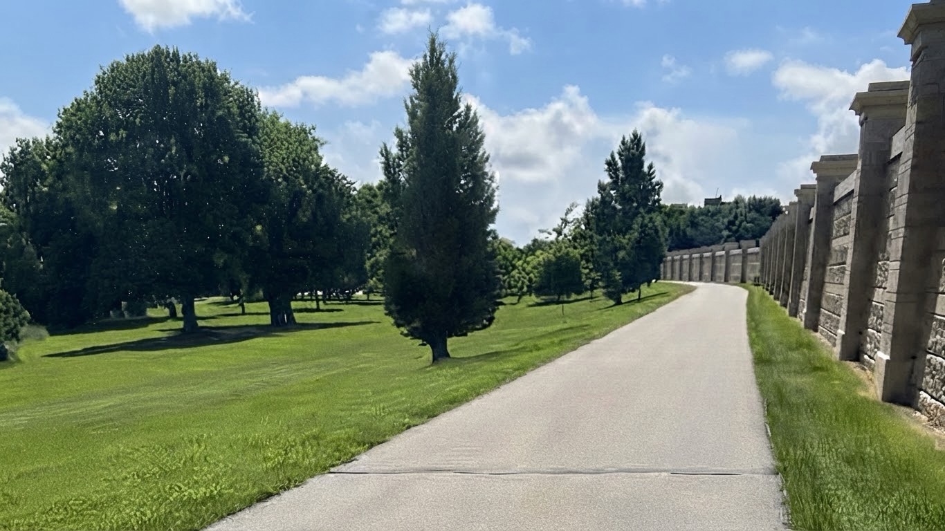 Paved Greenway path in Grand Forks running between the concrete flood wall and open grass with mature trees, with the post title The Grand Forks Greenway Was Never Supposed To Be a Park.