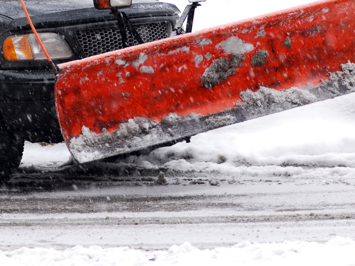 Orange snow plow on the front of a dark pickup truck in falling snow.